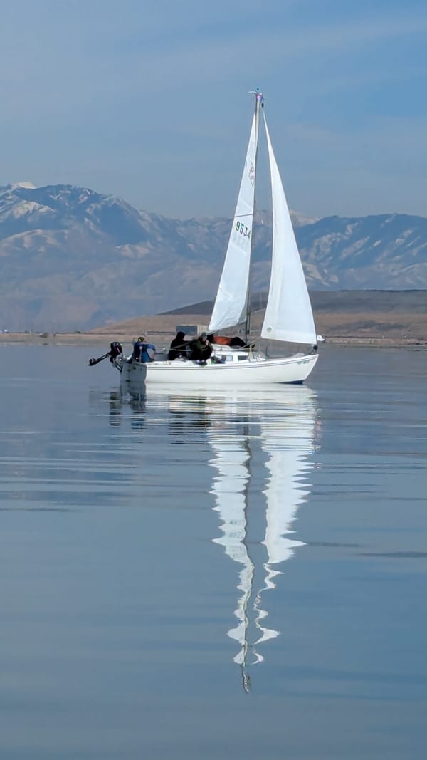 A sailboat sailing over calm waters.  The water reflects the sails with soft waves rippling the reflection of the sails.